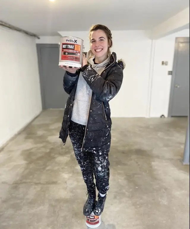 A esposa do Vini segurando uma lata de tinta de piso numa garagem